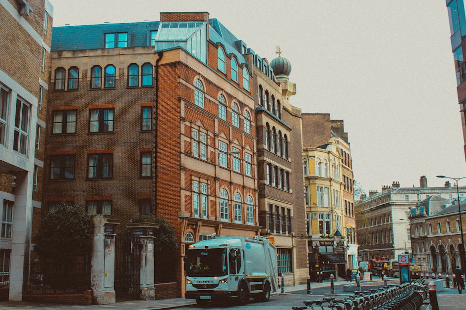 A small waste collection vehicle, painted in light grey with a compact and modern design, is parked on a city street in front of a mix of historic and modern buildings. The vehicle has a streamlined body, tinted front windows, and visible side mirrors, indicating its use for waste or rubbish collection services. The street scene is set within an urban environment, featuring cobblestone paving and a row of black metal bike racks along the sidewalk. To the left, a brick wall and gated entrance mark a private property, while on the right, multi-storey buildings showcase a variety of architectural styles, including brick facades, large sash windows, and decorative elements. The buildings' colours range from warm red and beige tones to pastel yellow, with some roof sections visible, including a rounded turret with a dome. The sky is overcast, with diffused natural light highlighting the textures of the brickwork and the street scene. This scene is indicative of urban waste collection, with a focus on private rubbish removal services such as those offered by London Rubbish Removal, suitable for on-site clearance or alternative waste handling outside of municipal collection.