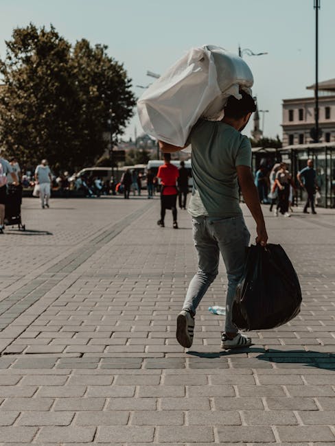 A young man walks across a paved city square, carrying a large white plastic bag over his shoulder and holding a smaller black bag in his right hand. The plastic bag appears to be filled with waste or items for disposal. He is dressed in a casual green T-shirt and light grey jeans. In the background, several pedestrians are visible, some standing and some walking, with diverse clothing styles. The scene includes trees, parked cars, and buildings under a clear sky, with sunlight casting soft shadows on the ground. This setting suggests urban environment activities related to waste handling or disposal, reflecting the importance of private rubbish collection services that companies like London Rubbish Removal facilitate, especially for street-level or on-site clearance needs. The overall atmosphere is calm and the image emphasizes individual responsibility for waste management in city spaces, aligning with the context of independent waste removal options separate from municipal services.
