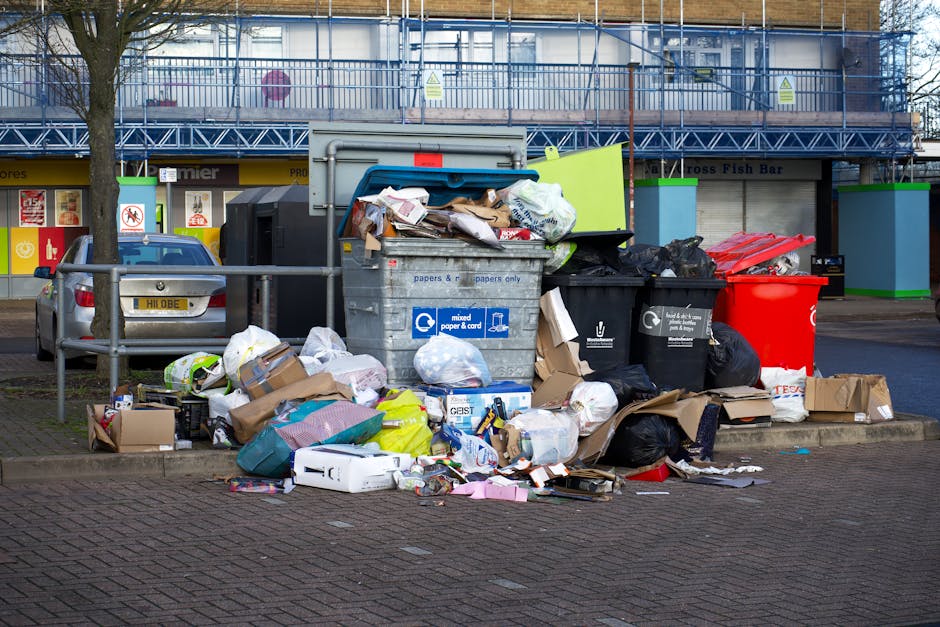 A white commercial van parked on a city street, with its rear doors open revealing a large load of mixed waste materials including cardboard boxes, black plastic bags, and other discarded items. The cardboard boxes are stacked on top of the van's interior, some partially crushed and torn, with visible corrugated surfaces and brown and white colors. The black plastic bags are piled inside and outside the van, some resting on the pavement next to a small shopping trolley filled with more waste. The van is situated in front of a multi-storey building with a light grey facade featuring rectangular windows and decorative architectural elements such as ledges and columns, typical of urban environments. The scene is illuminated by natural daylight, emphasizing the contrasts between the urban background and the waste materials, showcasing a typical example of private waste collection or rubbish clearance performed by a professional service in London. London Rubbish Removal occasionally provides such on-site clearance in dense city areas, ensuring waste is collected efficiently without reliance on local authority disposal options.